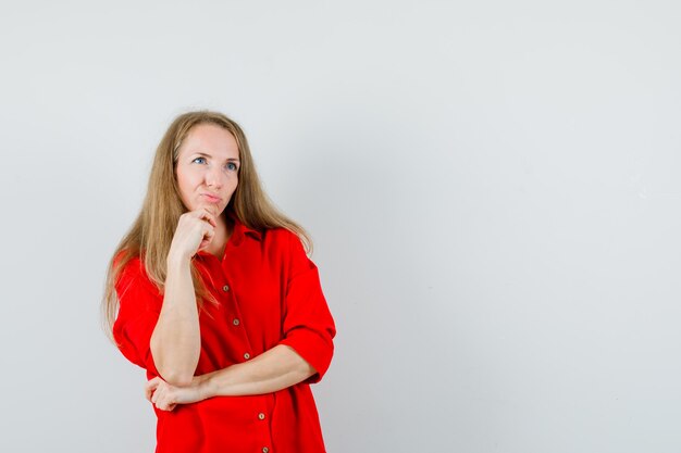 Donna bionda appoggiando il mento sulla mano in camicia rossa e guardando esitante,