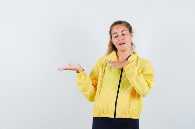 Donna bionda che allunga le mani come tenendo qualcosa di immaginario in bomber giallo e pantaloni neri e guardando concentrato