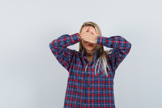 Donna bionda che copre gli occhi con entrambe le mani in camicia a quadri e guardando serio, vista frontale.