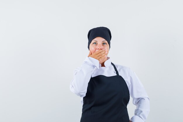 Donna bionda che copre la bocca con la mano in uniforme nera del cuoco e sembra carina