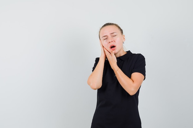 Donna bionda che finge di soffrire di mal di denti in maglietta nera e sembra esausta