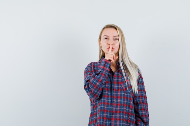 Donna bionda che mette il dito indice sulla bocca e mostra il gesto di silenzio in camicia a quadri e sembra attraente, vista frontale.