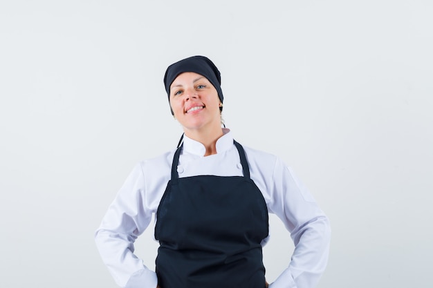 Donna bionda che mette le mani sulla vita, che propone alla macchina fotografica in uniforme nera del cuoco e che sembra carina. vista frontale.