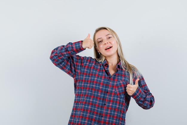 Donna bionda che mostra i doppi pollici in su in camicia controllata e che sembra felice, vista frontale.