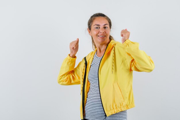 Donna bionda che mostra il gesto del vincitore in bomber giallo e camicia a righe e sembra carina