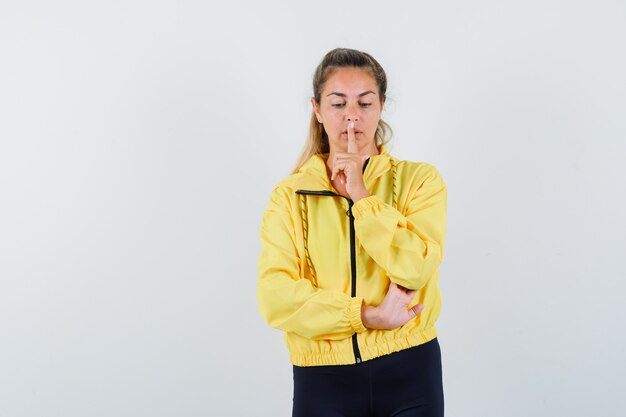 Donna bionda che mostra il gesto di silenzio in bomber giallo e pantaloni neri e guardando concentrato