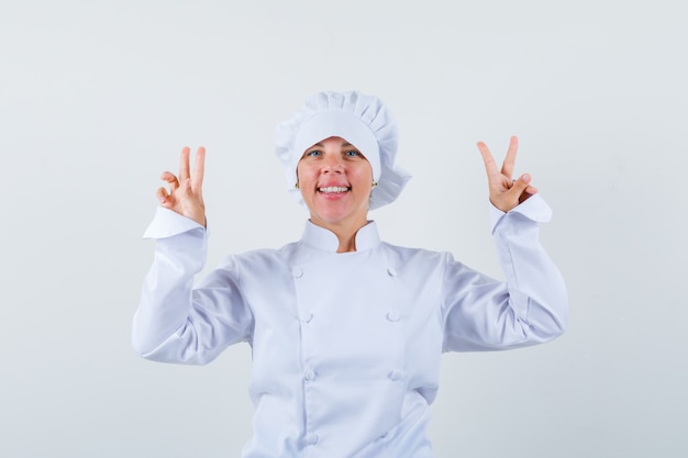 Donna bionda che mostra segni di pace con entrambe le mani in uniforme bianca del cuoco e che sembra carina