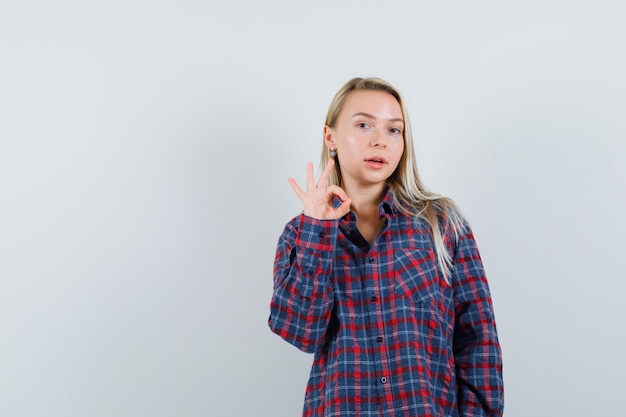 Donna bionda che mostra segno giusto in camicia a quadri e sembra attraente, vista frontale.