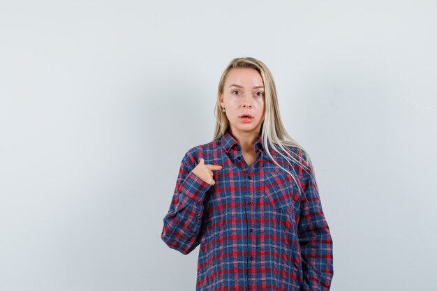 Donna bionda che punta a se stessa in camicia a quadri e che sembra sorpresa. vista frontale.