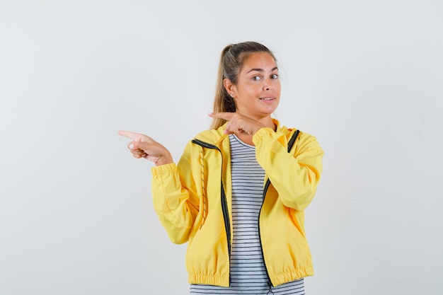 Donna bionda che punta a sinistra con il dito indice in bomber giallo e camicia a righe e sembra carina