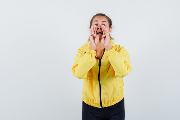 Donna bionda che tiene le mani vicino alla bocca come chiamare qualcuno in bomber giallo e pantaloni neri e che sembra felice