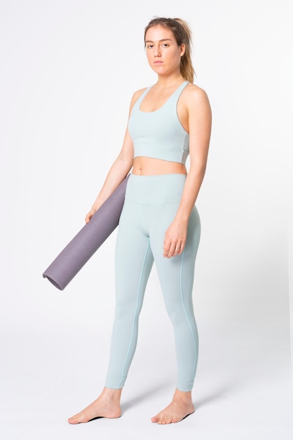 Donna bionda che tiene un tappetino da yoga in reggiseno sportivo blu e leggings a corpo intero