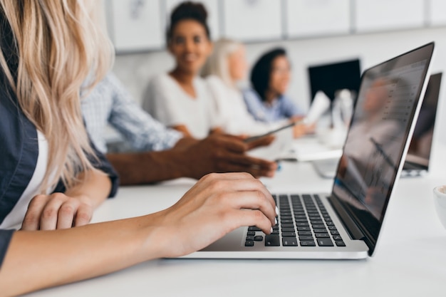 Donna bionda con acconciatura elegante che digita il testo sulla tastiera in ufficio. Ritratto dell'interno di dipendenti internazionali con segretaria utilizzando laptop.