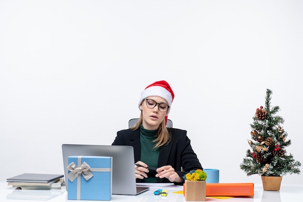 Donna bionda confusa con un cappello di Babbo Natale seduto a un tavolo con un albero di Natale e un regalo su di esso e guardando qualcosa con attenzione in ufficio su priorità bassa bianca