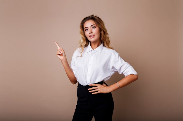 Donna bionda di affari che indica in su e che osserva sulla parete beige. Indossa abiti da lavoro eleganti. Copia spazio per il testo.