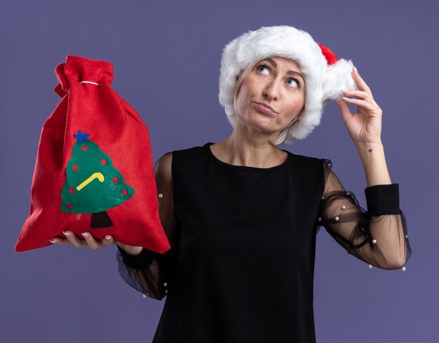 Donna bionda di mezza età confusa che porta il cappello di natale che tiene il cappello toccante del sacco di Natale che osserva in su isolato su fondo viola