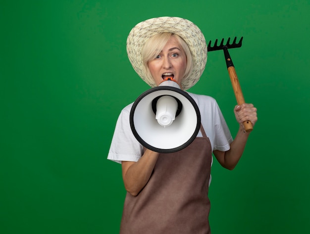 Donna bionda di mezza età del giardiniere in uniforme che indossa il cappello che alza il rastrello parlando dall'altoparlante isolato sulla parete verde con lo spazio della copia