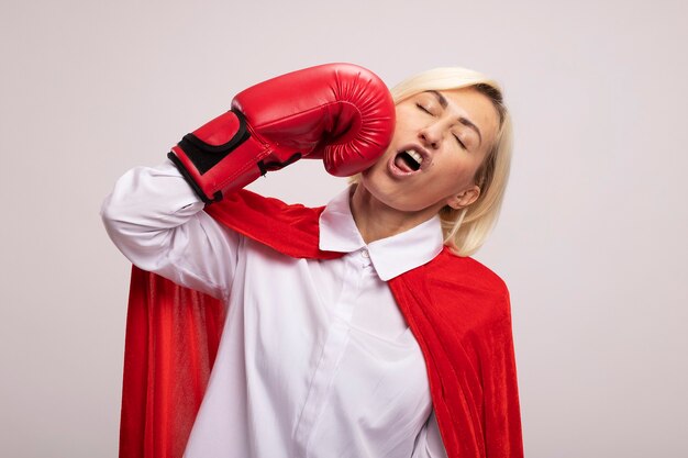 Donna bionda di mezza età del supereroe in mantello rosso che indossa guanti di scatola che si batte in faccia con gli occhi chiusi