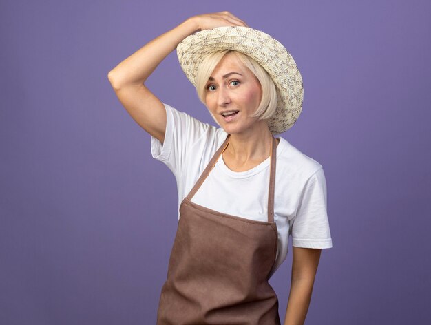 Donna bionda di mezza età impressionata del giardiniere in uniforme che indossa il cappello che mette la mano sul cappello