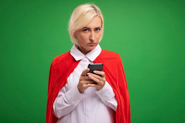 Donna bionda di mezza età seria del supereroe in mantello rosso che tiene il telefono cellulare