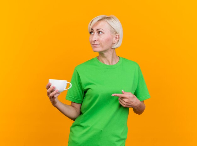 Donna bionda di mezza età sicura che tiene tazza di tè che osserva e che indica al lato isolato sulla parete gialla