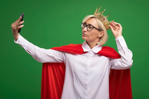 Donna bionda di mezza età sicura del supereroe in mantello rosso con gli occhiali e corona che afferra la corona che prende selfie isolato sulla parete verde