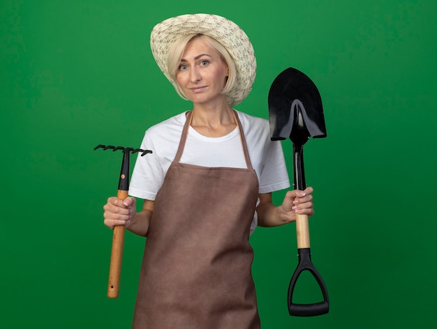 Donna bionda di mezza età soddisfatta del giardiniere in uniforme che indossa il cappello che tiene rastrello e vanga isolati sulla parete verde con lo spazio della copia