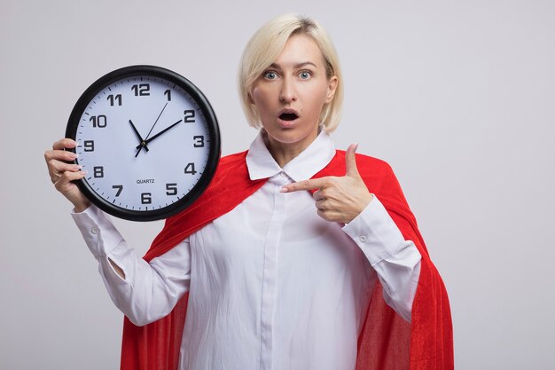Donna bionda di mezza età sorpresa del supereroe in mantello rosso che tiene e che indica l'orologio isolato sul muro bianco