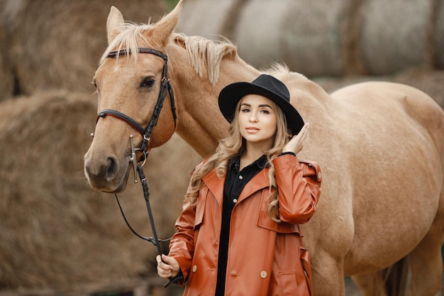 Donna bionda e cavallo marrone in piedi in una fattoria vicino a balle di fieno. Donna che indossa un abito nero, cappotto di pelle rossa e cappello. Donna che tocca il cavallo.