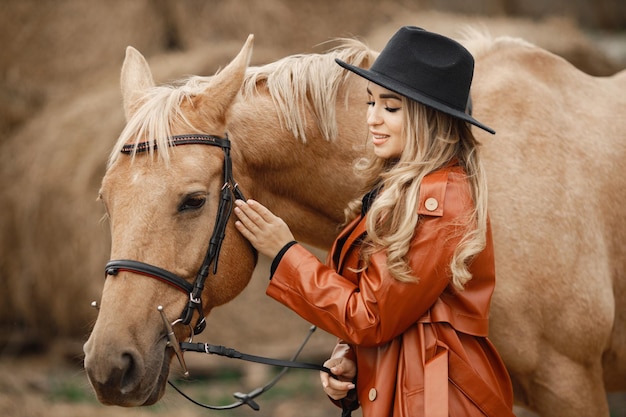Donna bionda e cavallo marrone in piedi in una fattoria vicino a balle di fieno. Donna che indossa un abito nero, cappotto di pelle rossa e cappello. Donna che tocca il cavallo.