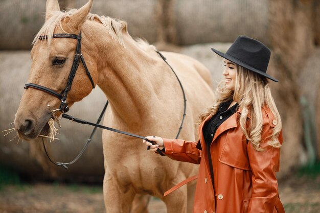 Donna bionda e cavallo marrone in piedi in una fattoria vicino a balle di fieno. Donna che indossa un abito nero, cappotto di pelle rossa e cappello. Donna che tocca il cavallo.