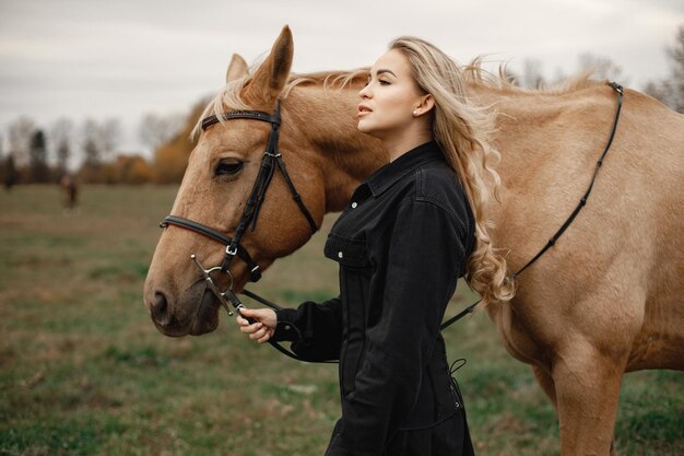 Donna bionda e cavallo marrone in piedi nel campo. Donna che indossa abiti neri. Donna che tocca il cavallo.