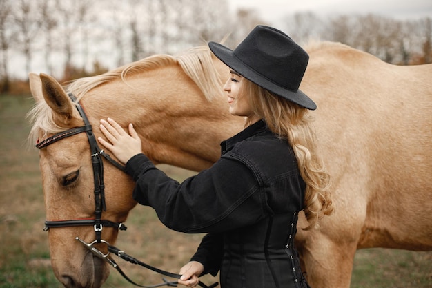 Donna bionda e cavallo marrone in piedi nel campo. Donna che indossa abiti neri e cappello. Donna che tocca il cavallo.