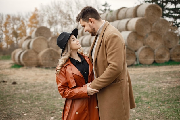 Donna bionda e uomo bruna che si abbracciano in una fattoria vicino alle balle di fieno. Donna che indossa abiti neri e cappotto di pelle rossa e uomo che indossa un cappotto marrone. Uomo e donna che toccano il cavallo