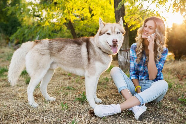 Donna bionda felice abbastanza giovane abbastanza sorridente che gioca con la razza del cane husky nel parco il giorno di estate soleggiato