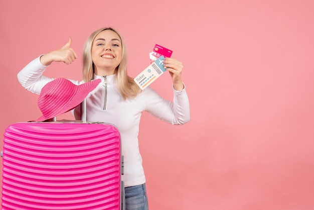 Donna bionda felice di vista frontale con la carta rosa della tenuta della valigia ed il biglietto aereo che danno i pollici in su