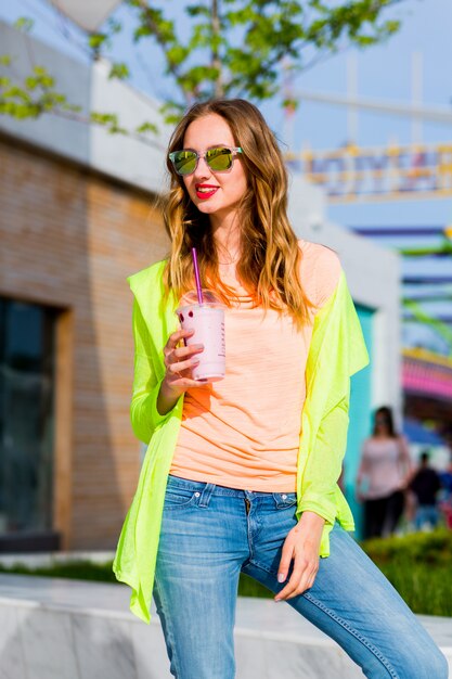 Donna bionda giovane hipster alla moda in occhiali da sole cool con cocktail in posa all'aperto