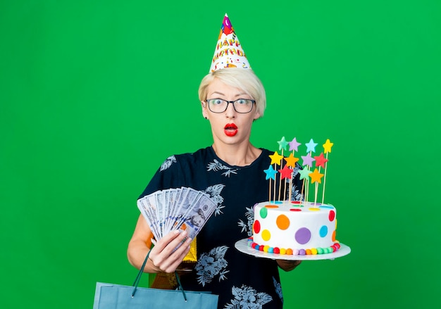 Donna bionda giovane sorpresa del partito con gli occhiali e berretto di compleanno che tiene la torta di compleanno con il contenitore di regalo dei soldi delle stelle e il sacchetto di carta che esamina la parte anteriore isolata sulla parete verde con lo spazio della copia