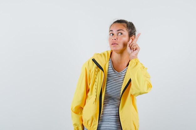 Donna bionda in bomber giallo e camicia a righe alzando il dito indice in gesto di eureka, distogliendo lo sguardo e guardando pensieroso