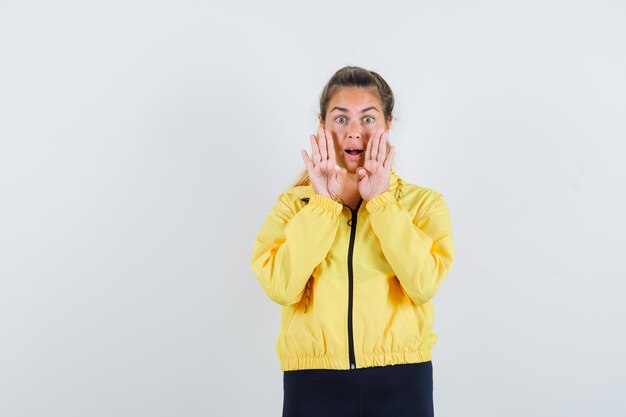 Donna bionda in bomber giallo e pantaloni neri che tengono le mani vicino alla bocca come chiamare qualcuno e che sembra felice