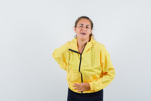 Donna bionda in bomber giallo e pantaloni neri, mettendo la mano sulla pancia, avendo mal di pancia e guardando esausto