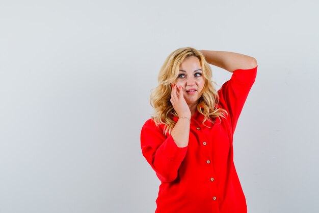 Donna bionda in camicetta rossa che tiene una mano dietro la testa, un'altra mano sulla guancia, che propone alla macchina fotografica e che sembra felice