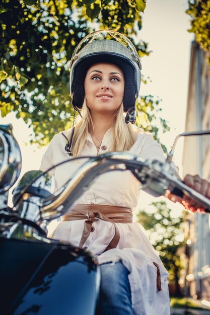 Donna bionda in casco moto seduto su scooter moto su foglie verdi e sole.