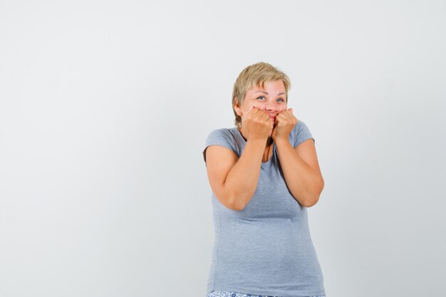 Donna bionda in maglietta azzurra che si morde i pugni, mette le mani sulle guance e sembra sorpresa, vista frontale.