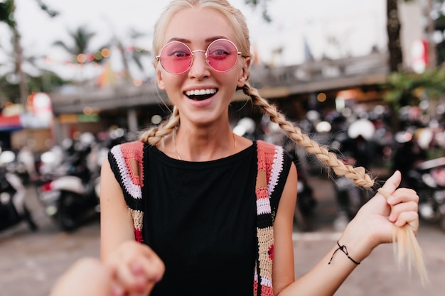 Donna bionda in occhiali da sole rosa in posa con un sorriso sorpreso. Donna che ride con le trecce che esprimono stupore sulla sfocatura dello sfondo della strada.