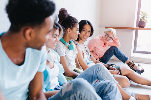 donna bionda in occhiali e camicia rosa seduta sul pavimento e guardando con interesse i suoi compagni di classe internazionali. Ritratto di studenti che si rilassano nel campus.