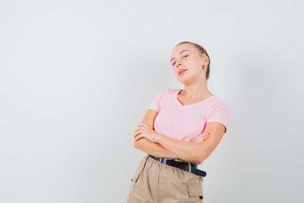 Donna bionda in piedi con le braccia incrociate in t-shirt, pantaloni e guardando fiducioso, vista frontale.