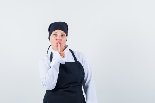 Donna bionda in uniforme nera del cuoco che mostra il gesto di silenzio e che sembra carina