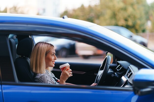 Donna bionda moderna che mangia un caffè per andare mentre si guida in città