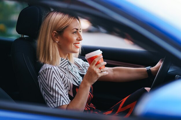 Donna bionda moderna che mangia un caffè per andare mentre si guida in città